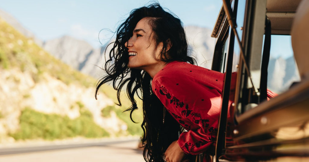 A close-up of a woman who has her head and upper body leaning outside of a car window. Her hair is blowing in the wind.