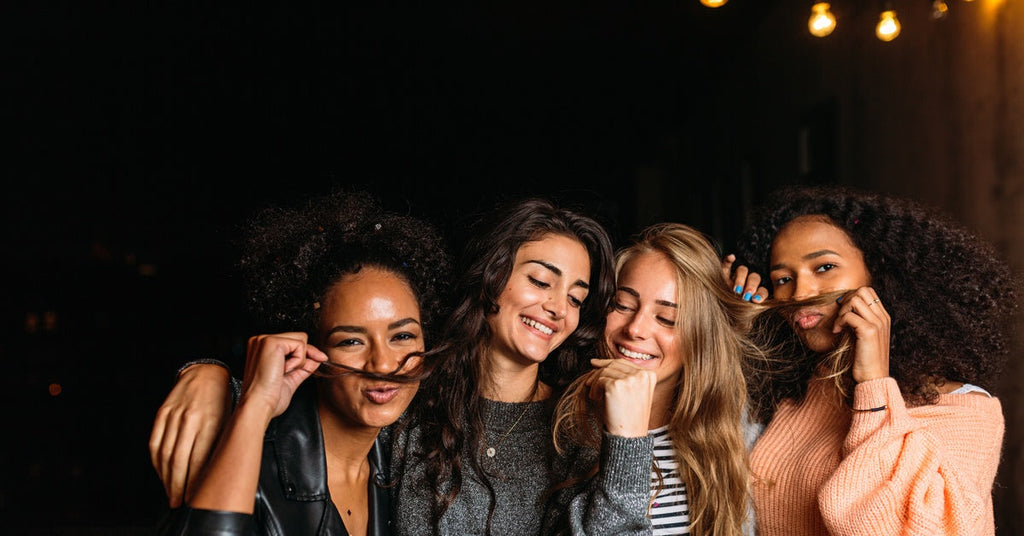Four young women are out having a girls' night. They're making silly faces and posing with their hair as mustaches.