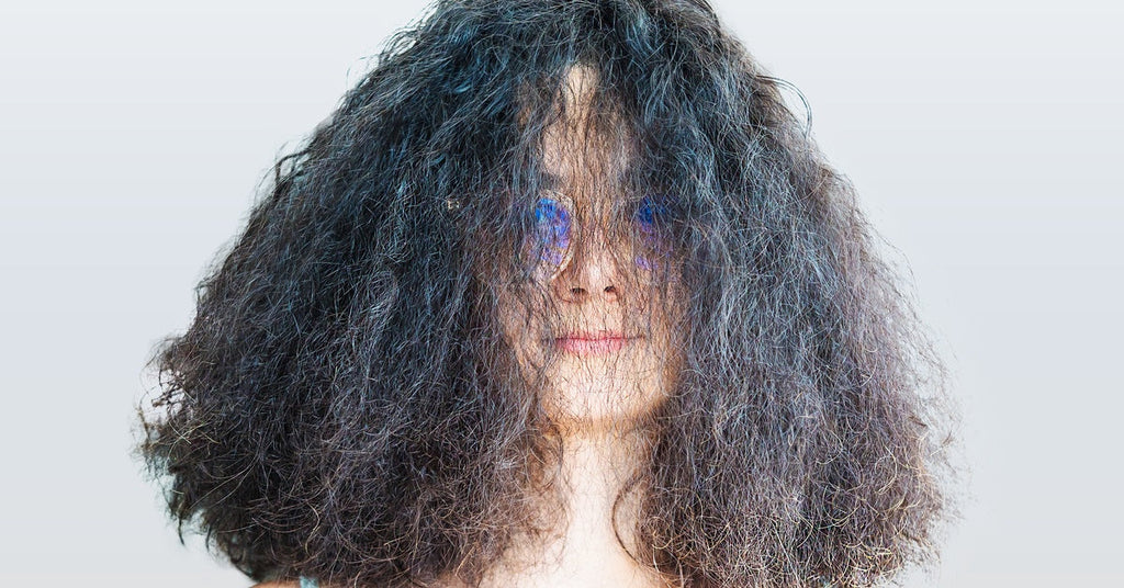 A portrait of a young woman with frizzy, shoulder-length hair covering her face. She wears glasses and has a neutral expression.