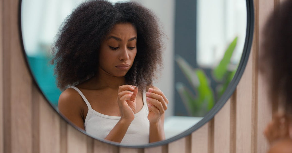 A young Black woman inspects the ends of her hair strands with a worried expression in front of her bathroom mirror.