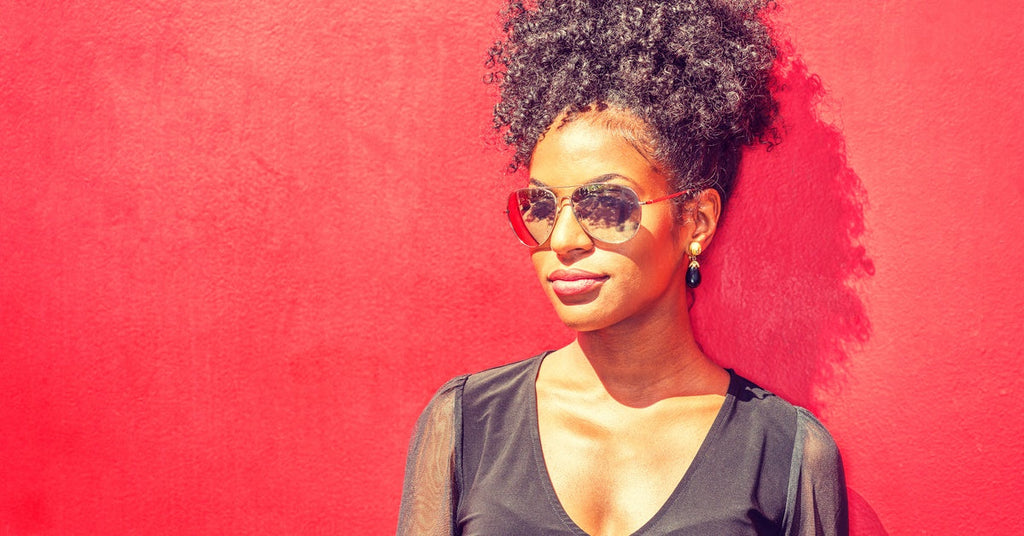 A young Black woman with curly hair styled in a tall bun is wearing sunglasses while standing against a red wall.