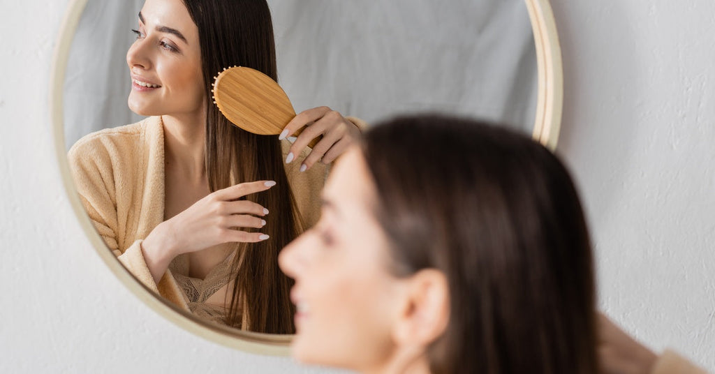 A cheerful woman uses a wooden brush on her straight, brunette hair in front of her bathroom mirror.
