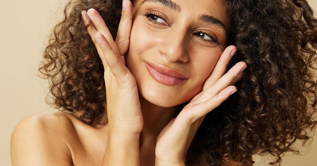 A close-up of a woman’s face as she poses with her hands on her cheeks and smiles. Her curly hair is worn down.
