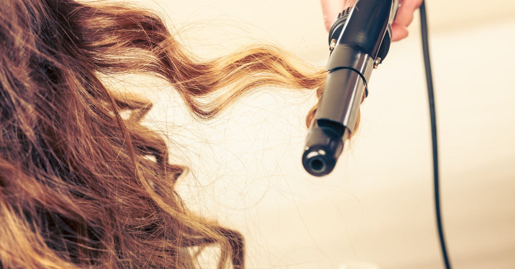 A person’s hand holding a curling iron, wrapping a strand of reddish-brown hair from a woman into soft curls.