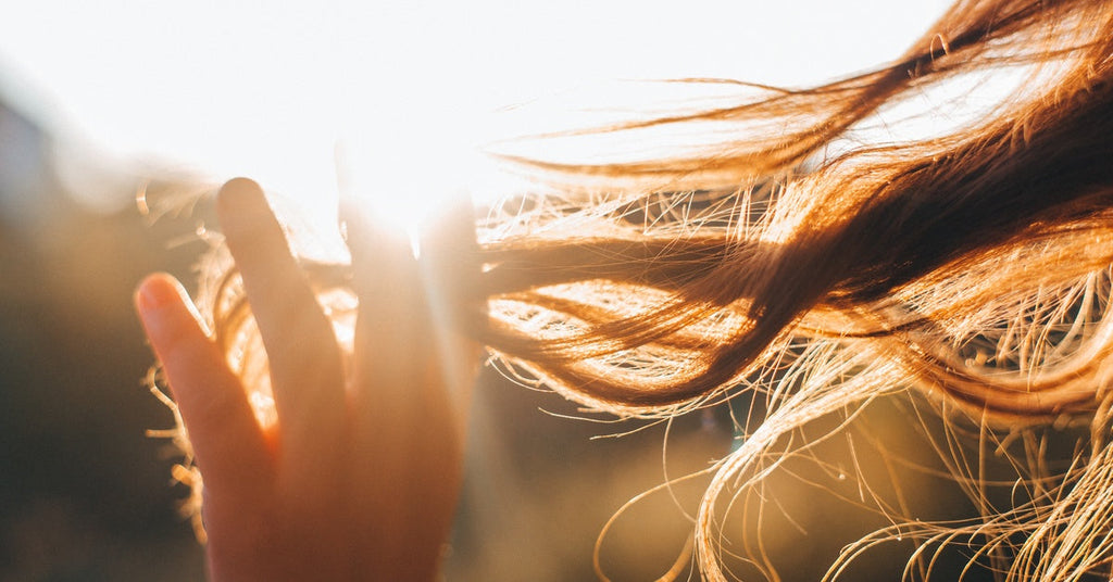 A hand touching a strand of hair blowing in the wind, with a bright beam of sunset illuminating the background.