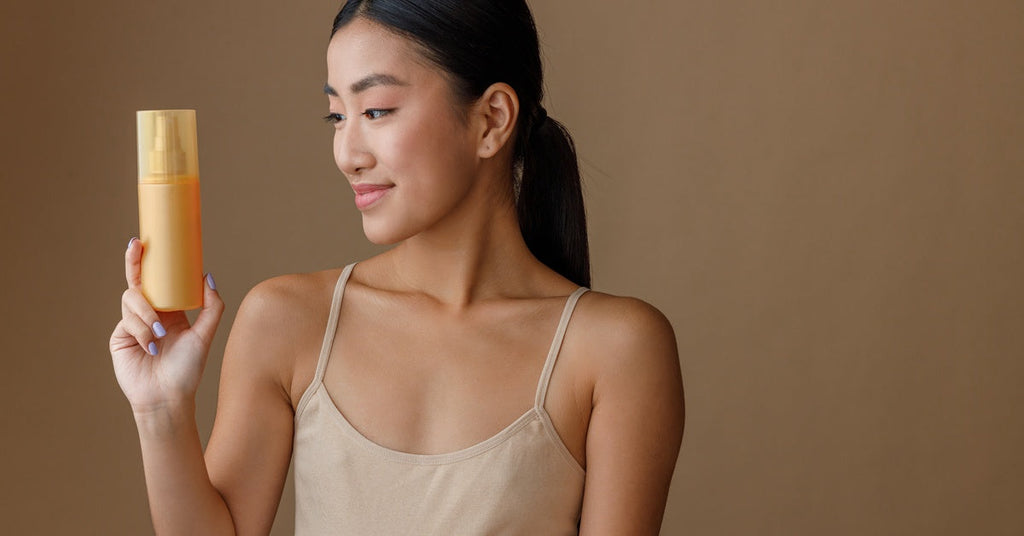 A young woman in a spaghetti strap top holding a translucent orange spray bottle and examining it against a brown background.
